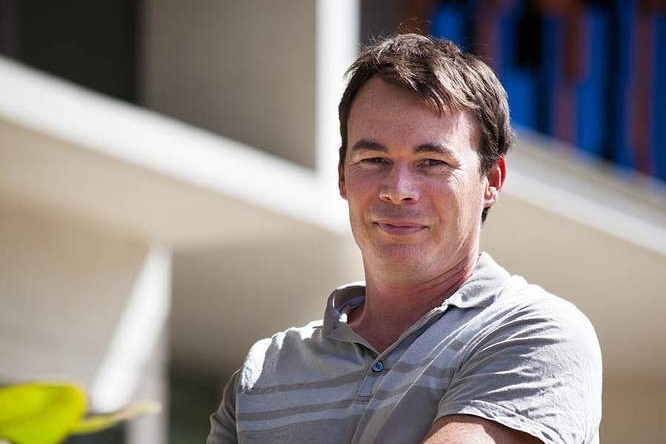 A caucasian man with short brown hair wearing a grey t-shirt standing outside on a sunny day with a building in the background