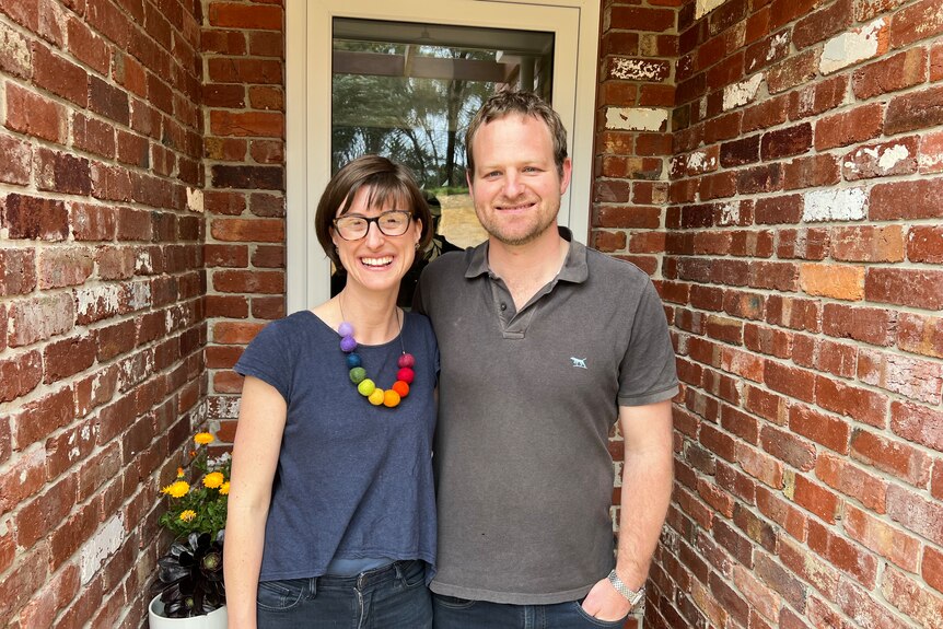 A young couple stand in a doorway with red bricks on either side. The woman has glasses and a colourful necklace