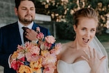 A man in a black suit wears a pink flower in his lapel and a woman in a white dress holds flowers and shows a ring on her finger