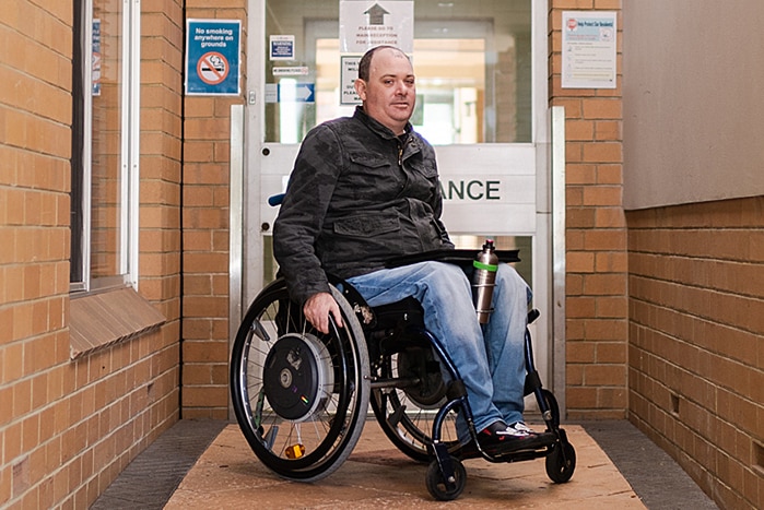 A man in a black shirt and jeans in a wheelchair in front of a building entrance.