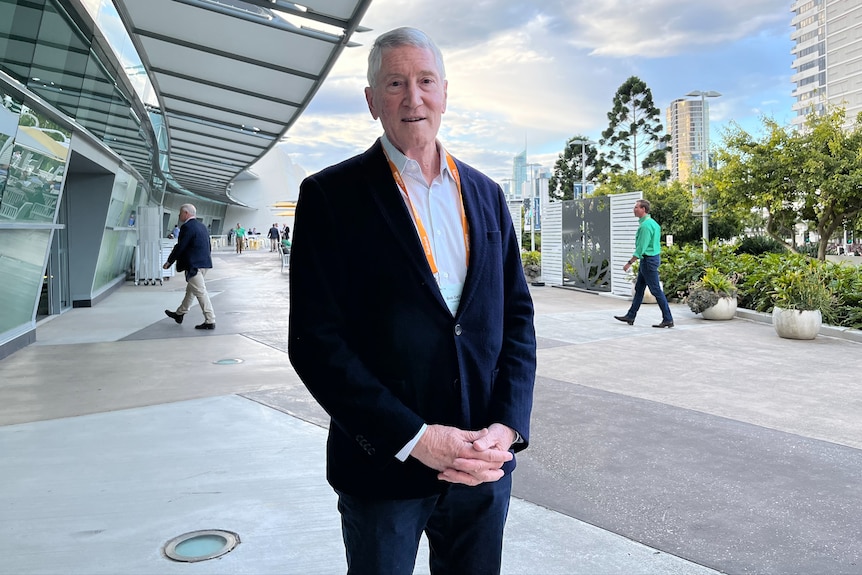 A grey-haired man stands holding his hands together.