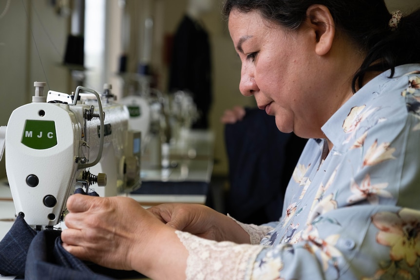 Amina wears a light blue top and feeds fabric through a sewing machine