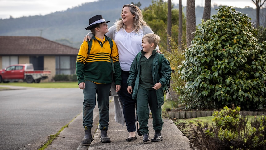 A mother and her two boys walk along the footpath on the way to school, smiling.