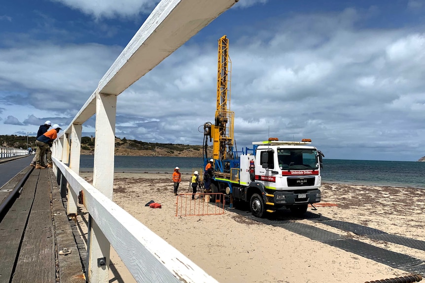 A truck with a drill on its tray top sits on a beach with workers surrounding it.