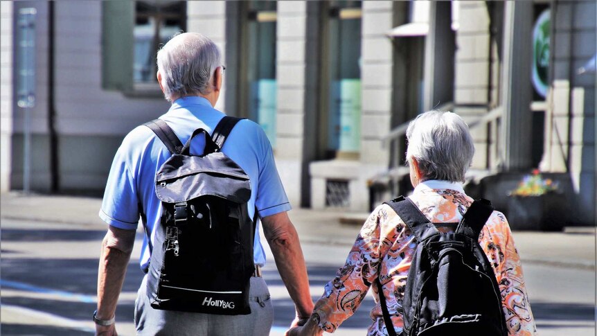 An older couple with grey hair and backpacks hold hands