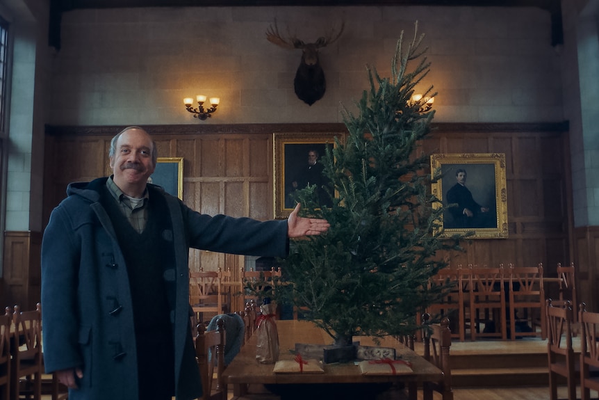 A film still of Paul Giamatti, with a sly smile, his arm gesturing towards a wonky Christmas tree on a table.