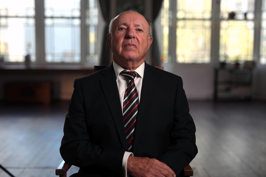 A man sits in a chair looking at the camera with a stern expression. Behind him are the windows of a large empty room.