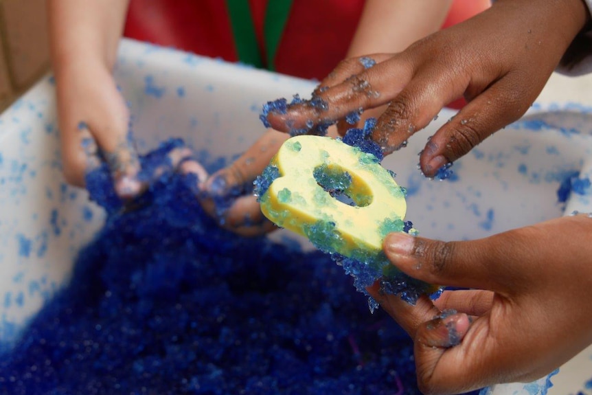 A close-up shot showing the hands of young children playing with a blue gel-like substance and giant letters at school.