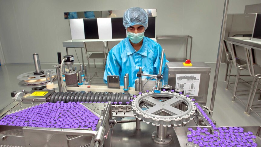 A man in PPE and a face mask examines a machine putting labels on purple medicine vials
