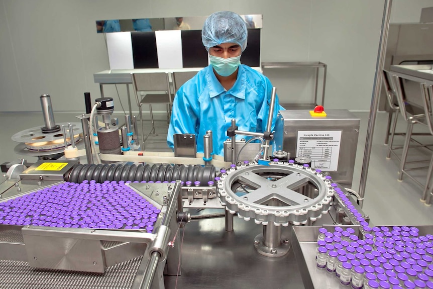 A man in PPE and a face mask examines a machine putting labels on purple medicine vials