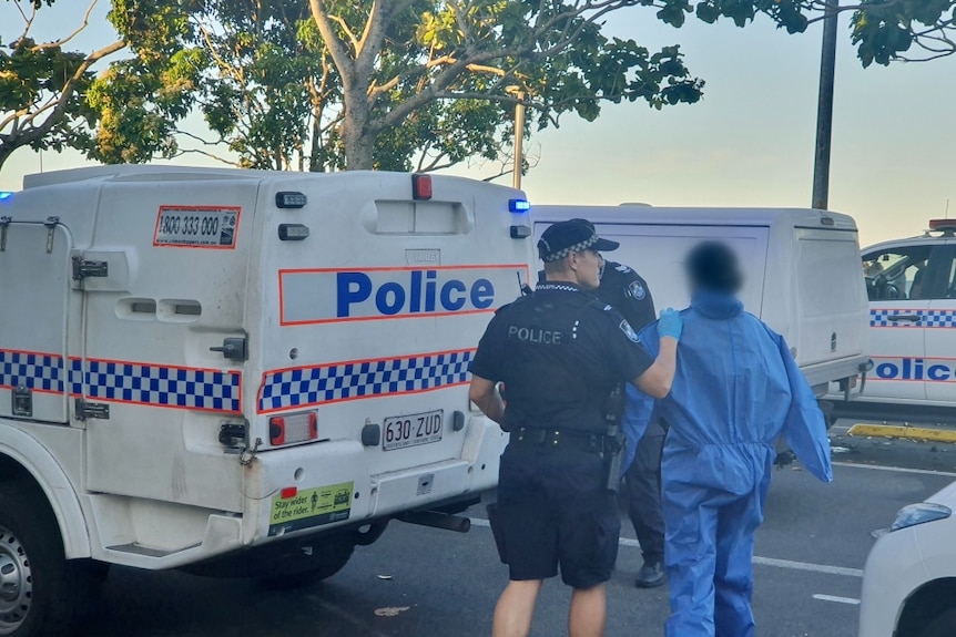 An unidentified person in disposable coveralls is seen from behind being escorted by a uniformed officer to a police vehicle