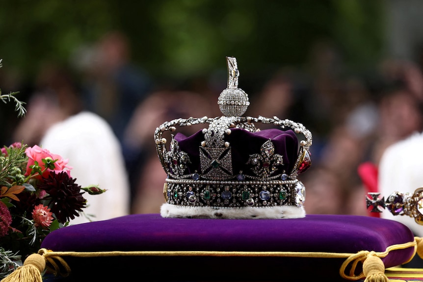 A purple and silver crown with crosses sits on a purple and gold cushion next to a bouquet of flowers.