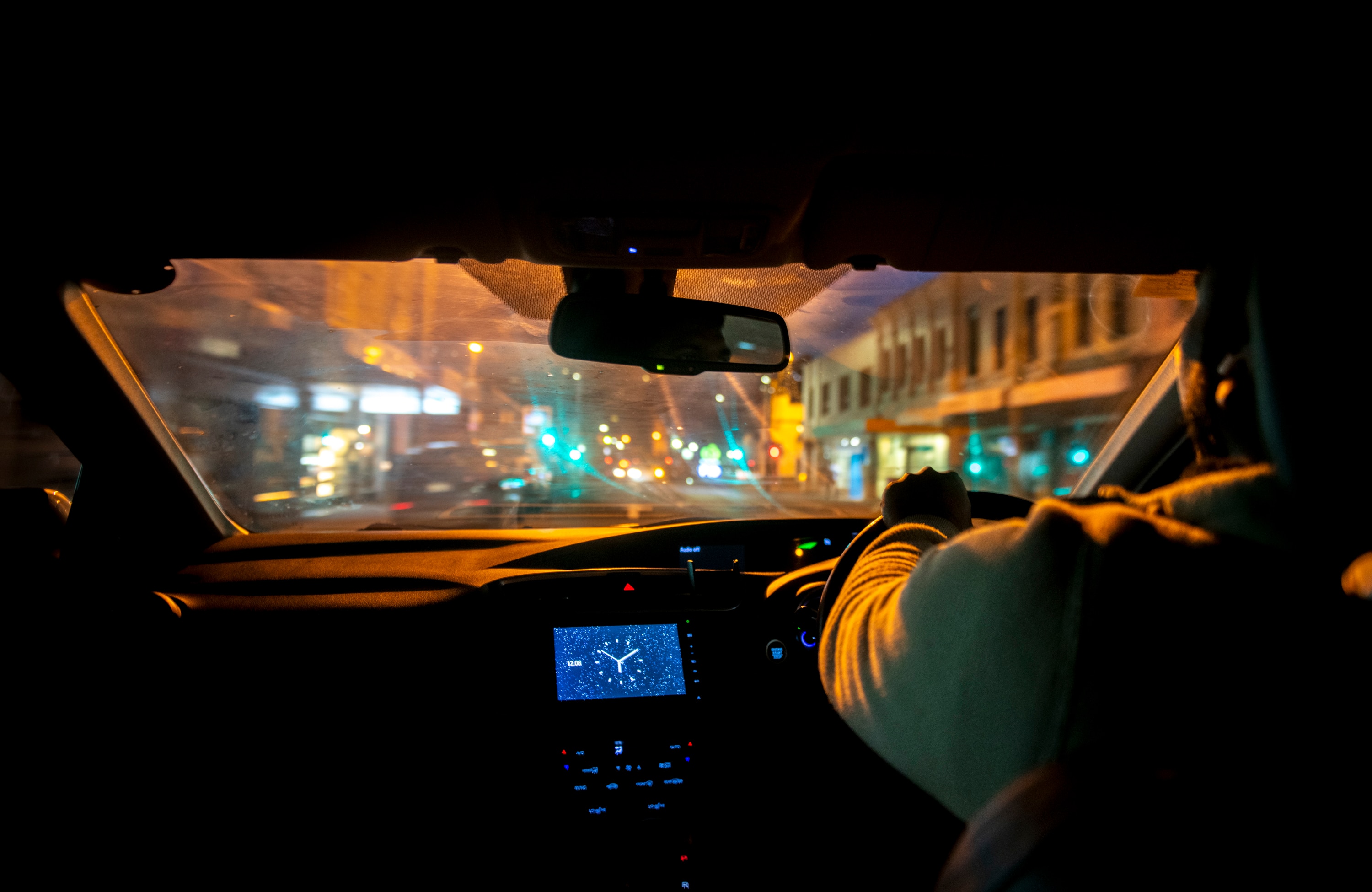 Gold street lamps and traffic lights glow through the windshield of a car driving passed buildings at night.
