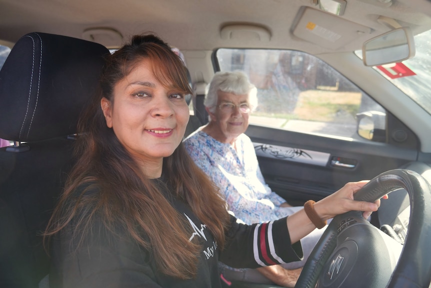 A woman sits behind the steering wheel of a car and holds it while a women sits in the passenger seat.