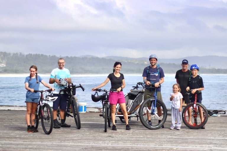 A group of adults and children standing with bikes and unicycles on a wharf by the ocean.