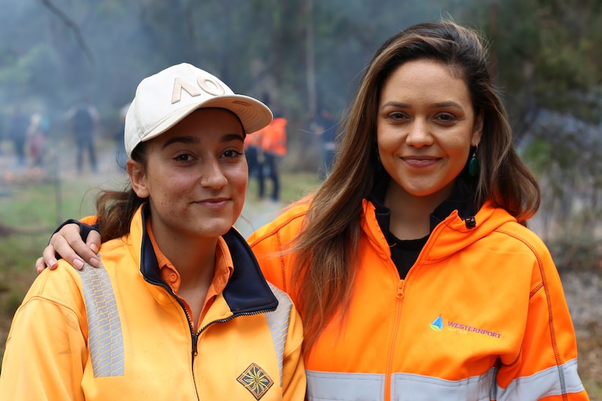 Two women stand beside one another in orange jackets in bushland as cultural burning occurs around them.