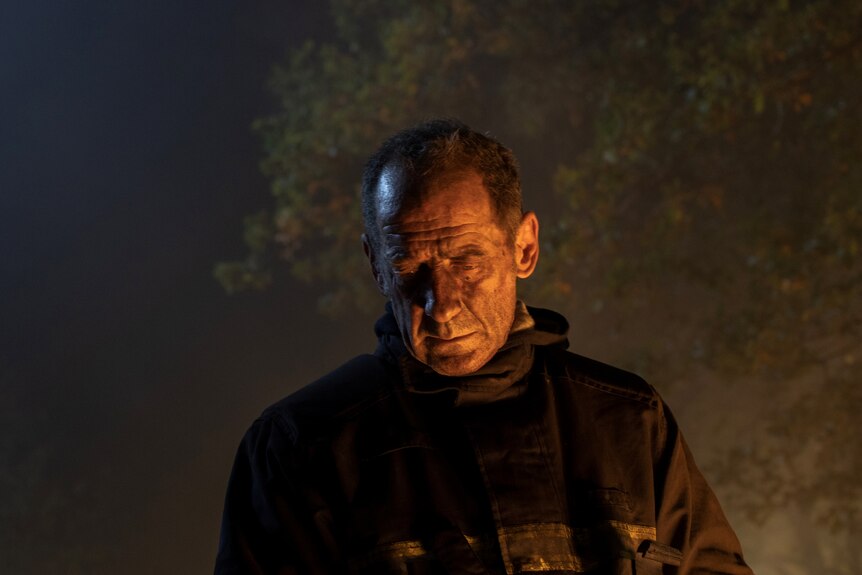 A man in his early 60s in dark fire fighter gear, ash on his face and smoke in the background
