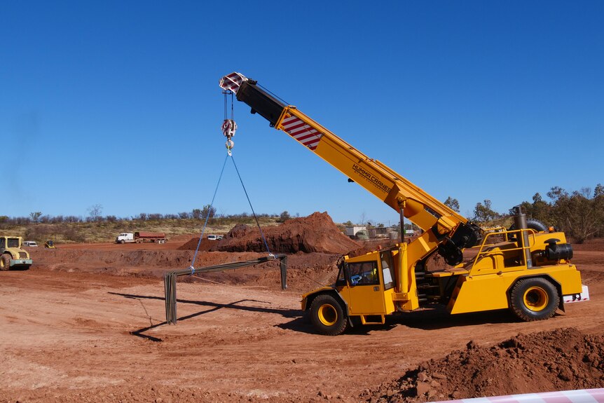 A big yellow crane truck carries some metal poles over a red earth works site under the blue Barkly sky.