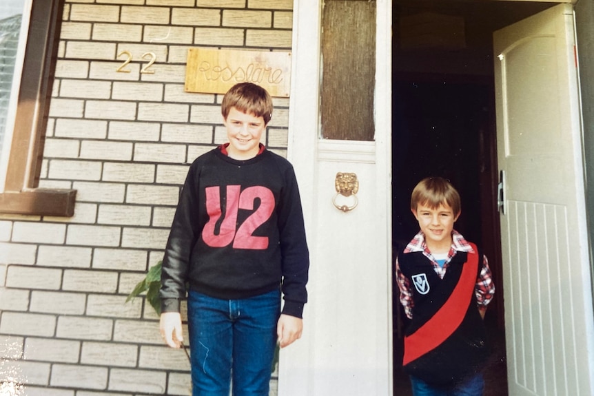 A tall, smiling boy stands in front of his house, with a younger boy beside him in a doorway.