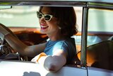 A woman in sunglasses and a blue dress leans out a vintage car
