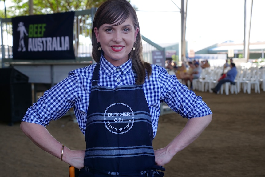 Una donna sorridente si mette le mani sui fianchi e indossa un grembiule scritto sopra "Butcher Girl Alison".