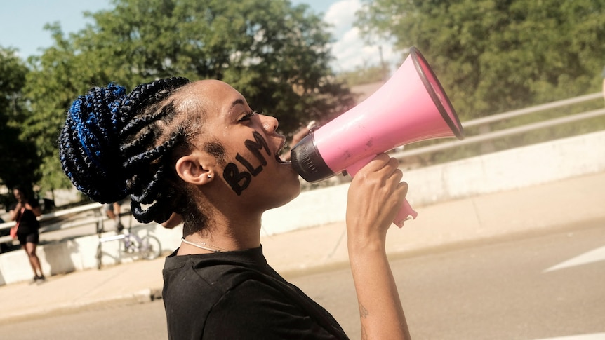 side profile of young Black woman holding megaphone to her mouth with "BLM" written on her cheek