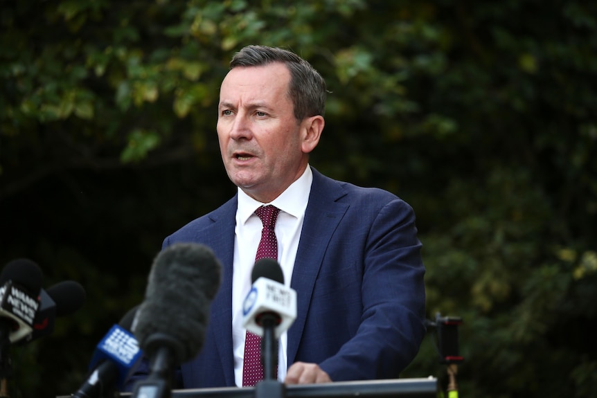 Mark McGowan wears a blue suit and purple tie, standing at a lectern with microphones speaking to the media.