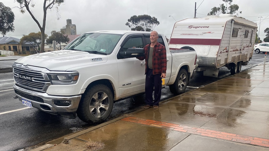 Man in plaid shirt stands next to white car hitched to a caravan, parked on the street.