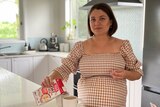 A pregnant woman with short dark hair, in a brown gingham op, standing in her kitchen