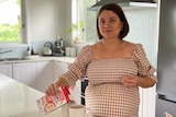 A pregnant woman with short dark hair, in a brown gingham op, standing in her kitchen