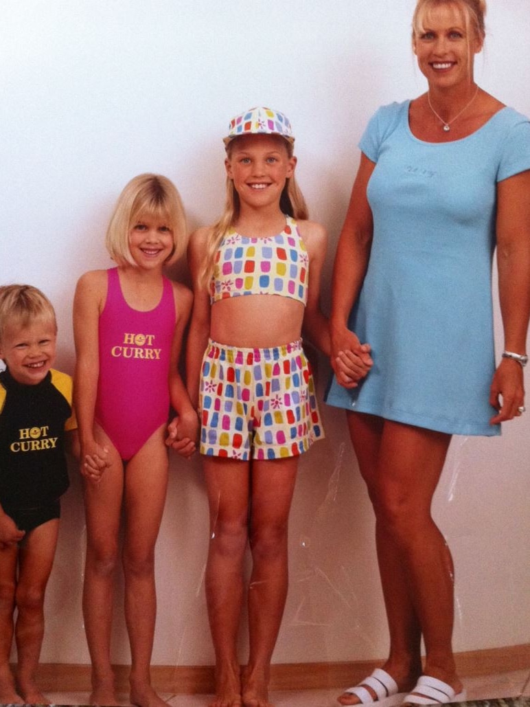 A blonde woman in blue summer dress stands with two daughters and a son who wear swimwear labelled Hot Curry