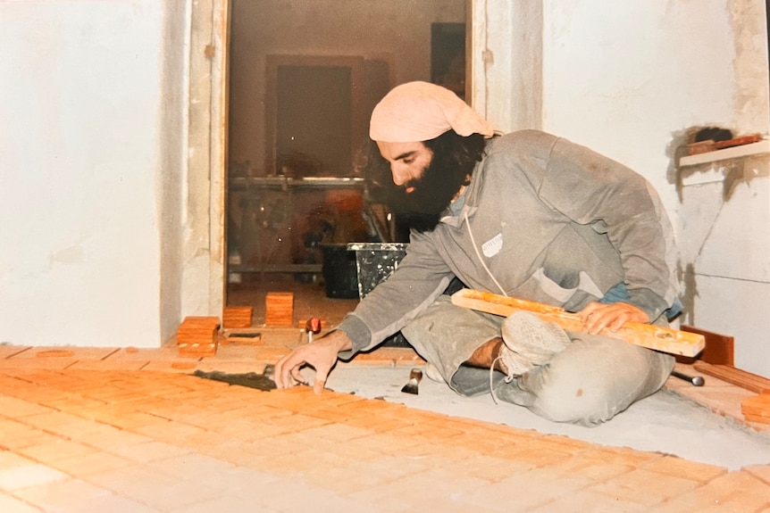 A man with a luscious, bushy beard meticulously slots in the next floor tile as he balances a spirit level on his knee as he