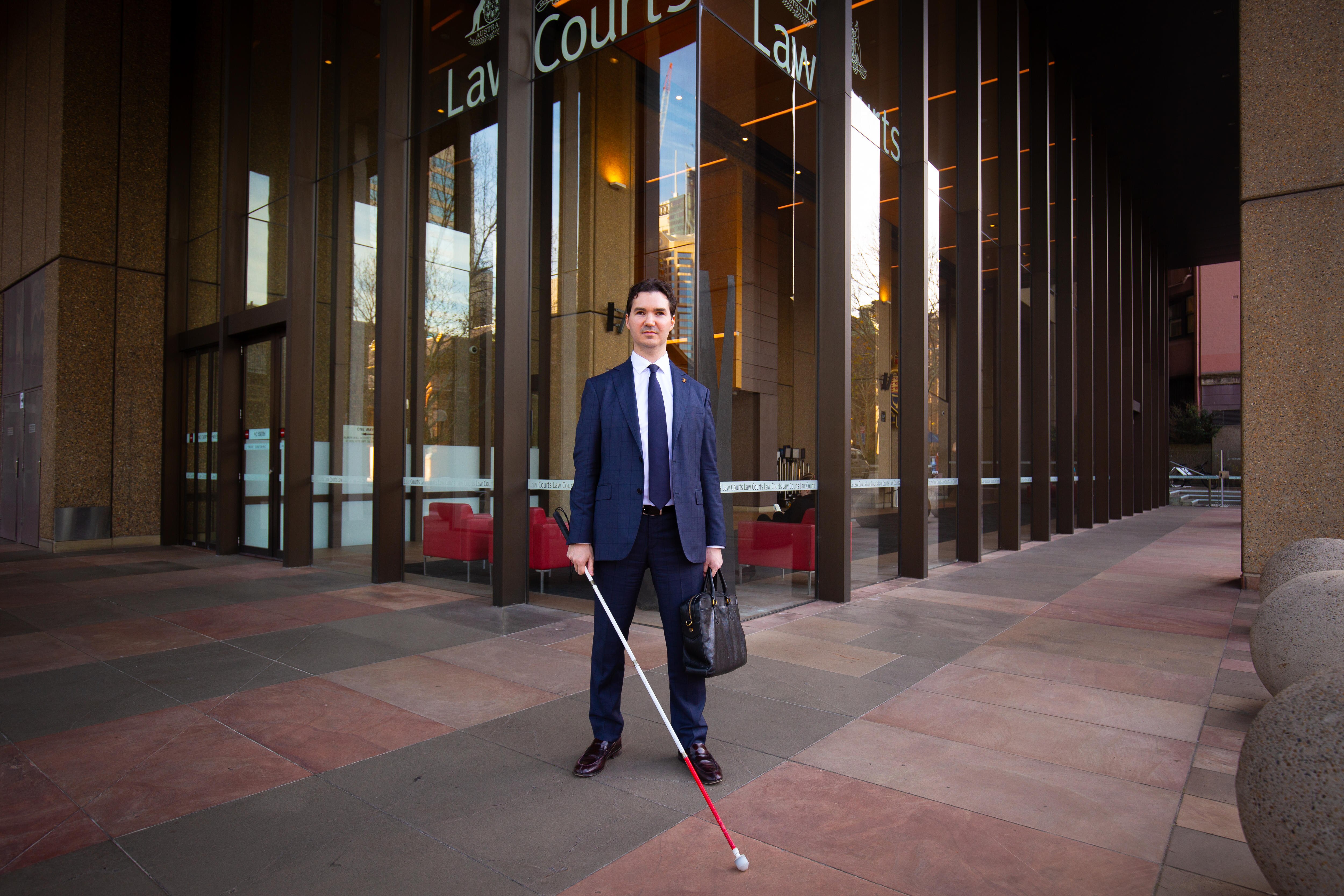 A white man with short hair and a blue suit standing outside a court building with a white cane and bag