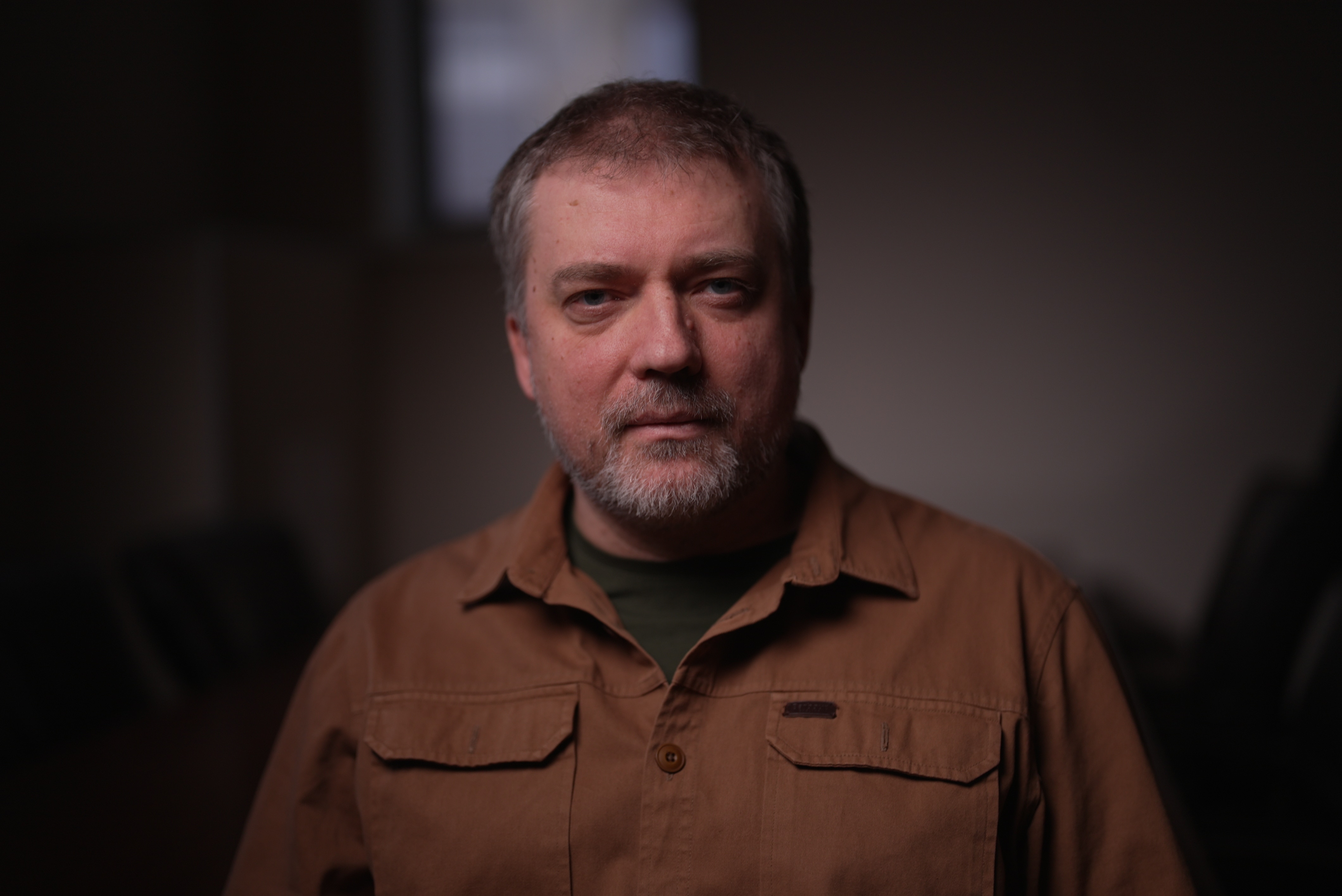 A man in a dark room looks at the camera with a neutral expression.