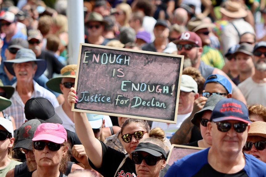 A large crowd in a town square, with one sign being held by a protester reading 'Enough is Enough'