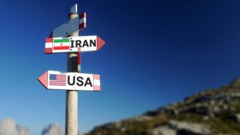On a sign post, one sign is facing right has 'Iran' written on it and the other has 'USA'