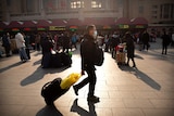 A man wearing a facemask drags his luggage in front of a train station.