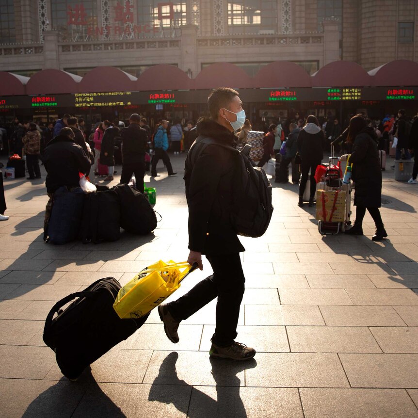 A man wearing a facemask drags his luggage in front of a train station.