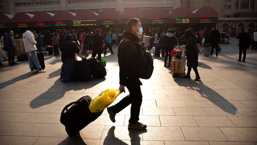 A man wearing a facemask drags his luggage in front of a train station.