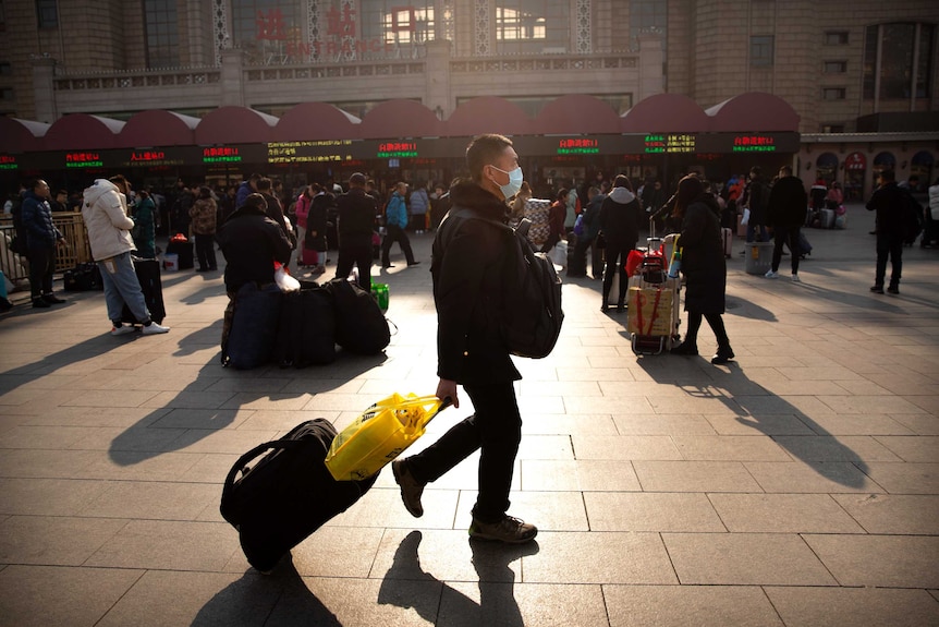 A man wearing a facemask drags his luggage in front of a train station.