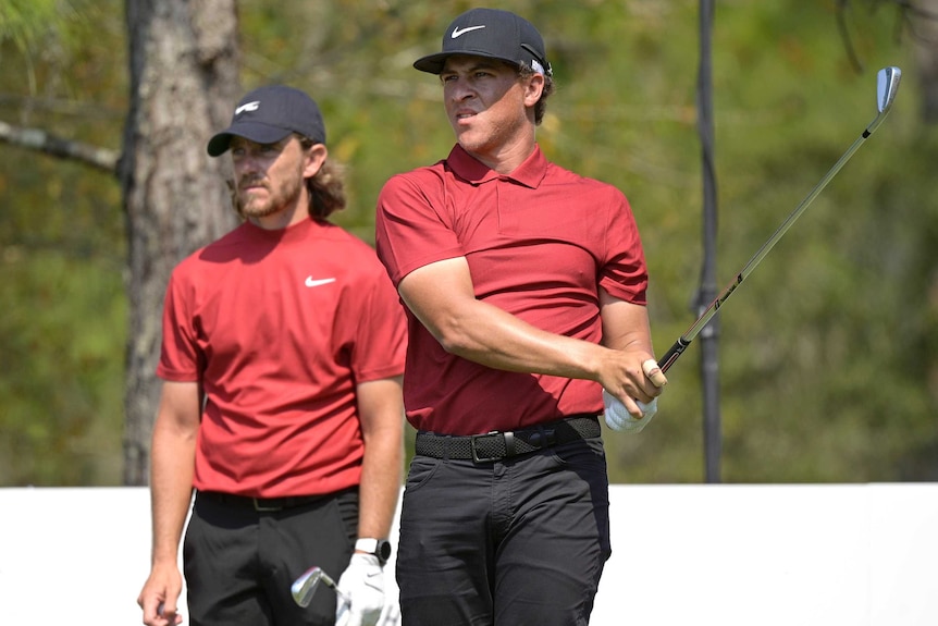Two male golfers wearing red shirts and black hats in the act of playing a shot