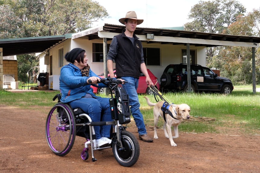 Woman in wheelchair travelling alongside a farmer and his guide dog walking past a house in a paddock.