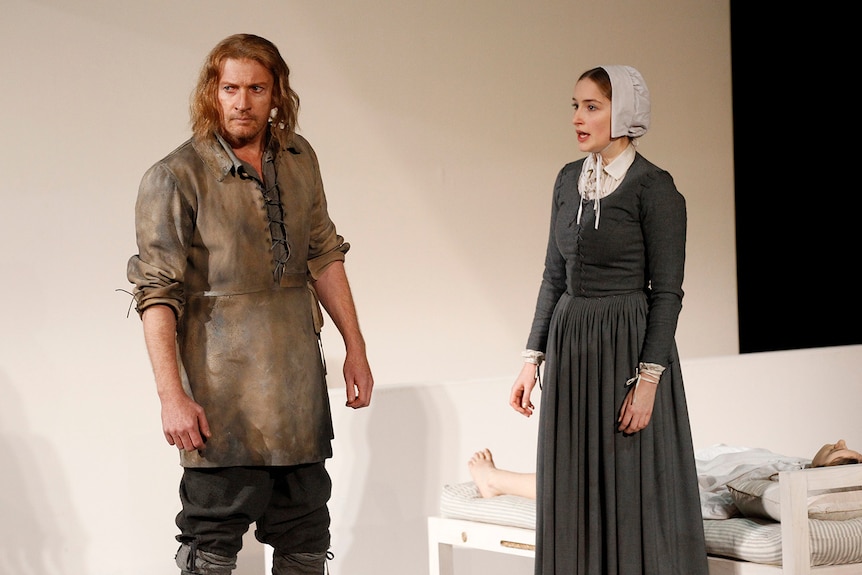 On stage, a middle-aged man looks stern. A woman speaks to him, another lies on a bed behind. They're in in early modern dress.