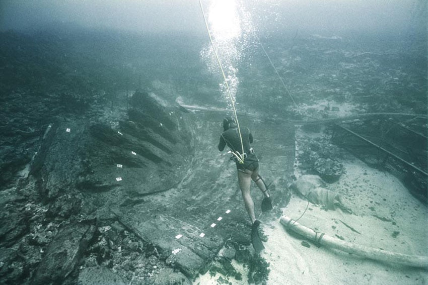 Man in black scuba wetsuit with rope around waist and section of curved timber ship hull protuding from ocean floor. 