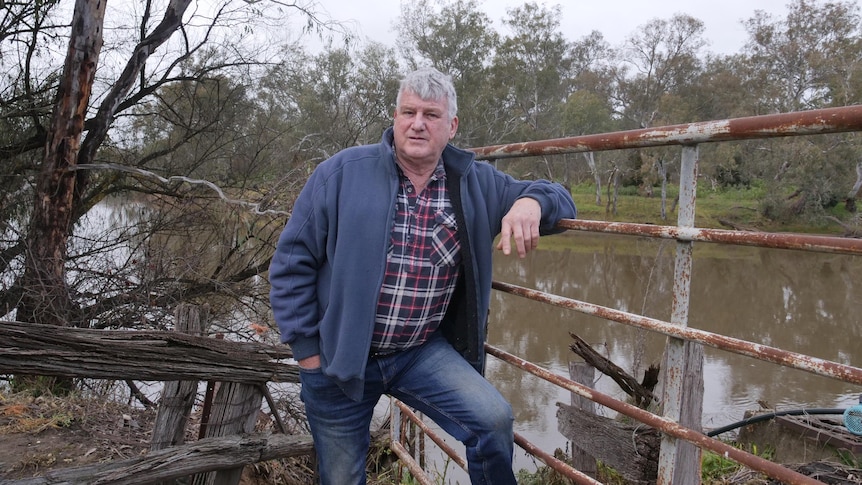 A man leans on a metal gate. He is wearing blue jeans, blue jacket and a red, white and blue striped shirt