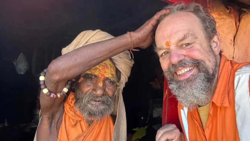Bearded man smiles as an Indian bearded man holds his hand on his head. Both wear orange, drapped clothing 