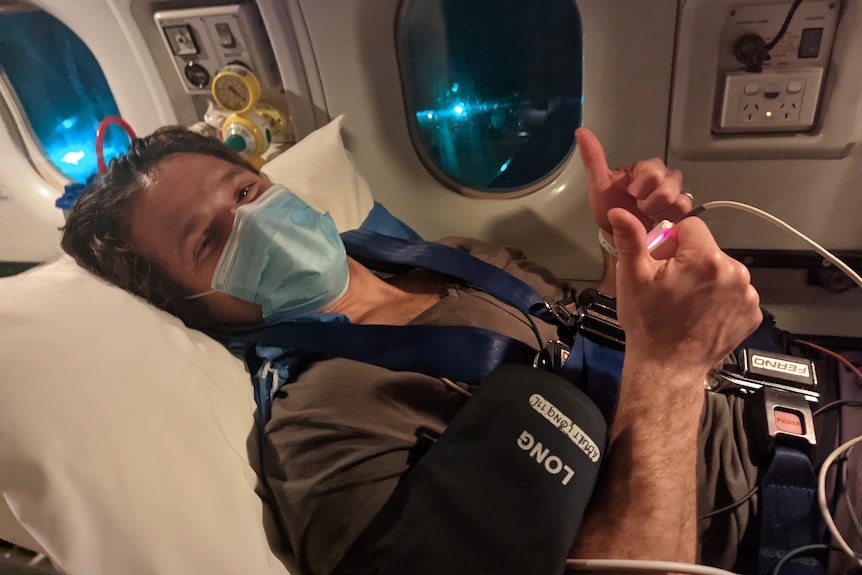 Smiling man lying strapped in an airplane wearing a mask gives thumbs up.