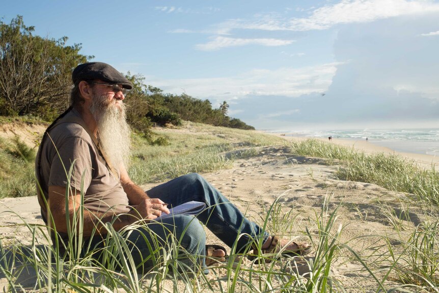 A man with a grey beard wearing a cap sits with a notebook on the sand dunes