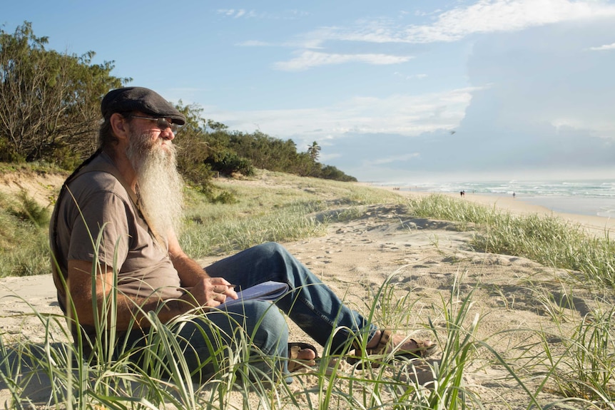 A man with a grey beard wearing a cap sits with a notebook on the sand dunes