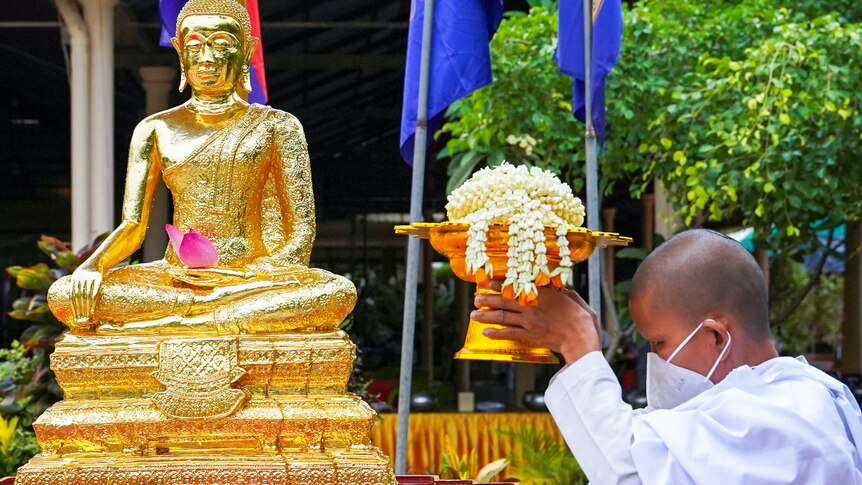 A Cambodian nun in a face mask holds a golden bowl filled with flowers next to a golden Buddha statue